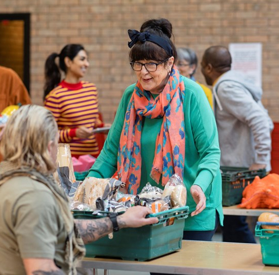 A woman wearing glasses, a black headband, and a colourful scarf hands a green crate of food to another person at a community food distribution event. Other volunteers and tables with food items are visible in the background.