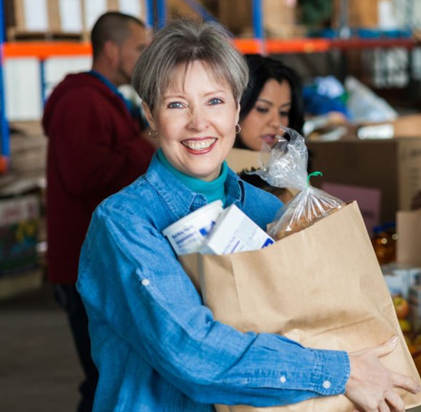 A woman holding a brown bag of groceries smiling directly at the camera