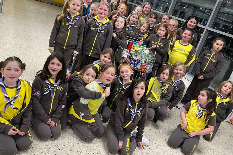 Group of Brownies smiling with their leaders and a trolley full of groceries indoors.